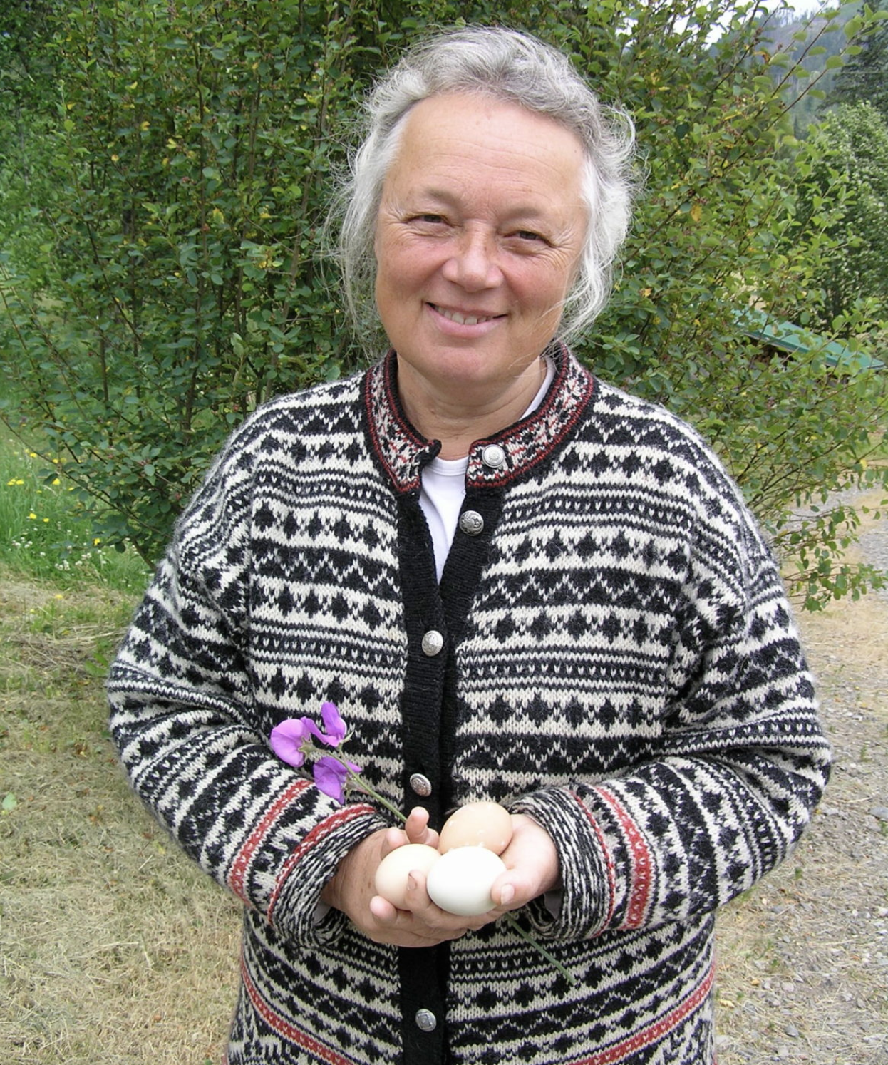 A woman with grey hair smiling outdoors, holding four eggs and a purple flower, wearing a patterned sweater.