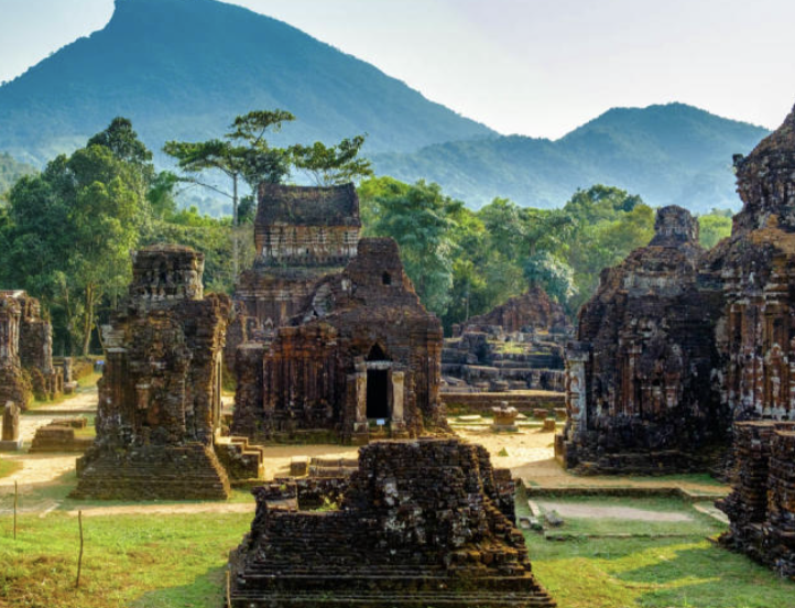 Ancient brick stupa with a single tall pillar topped by a small sculpture, surrounded by trees and ruins.