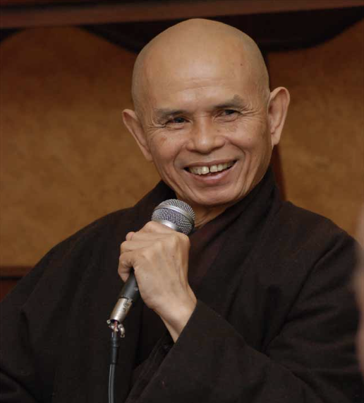 A smiling bald man dressed in traditional black robes, holding a microphone, speaking at an event.