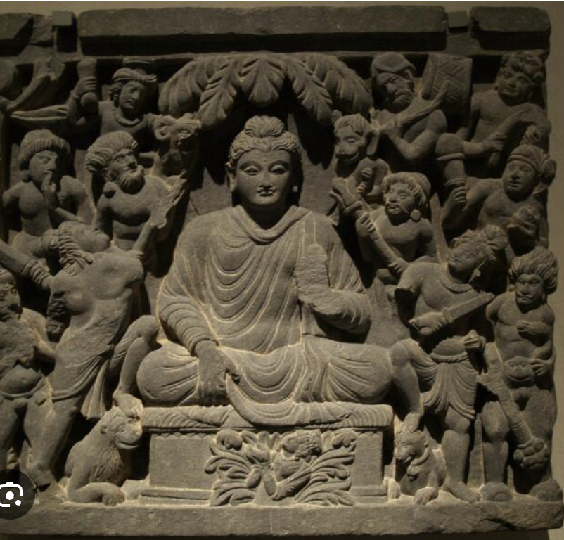 Stone sculpture of a seated Buddhist figure surrounded by smaller figures, animals, and decorative elements.