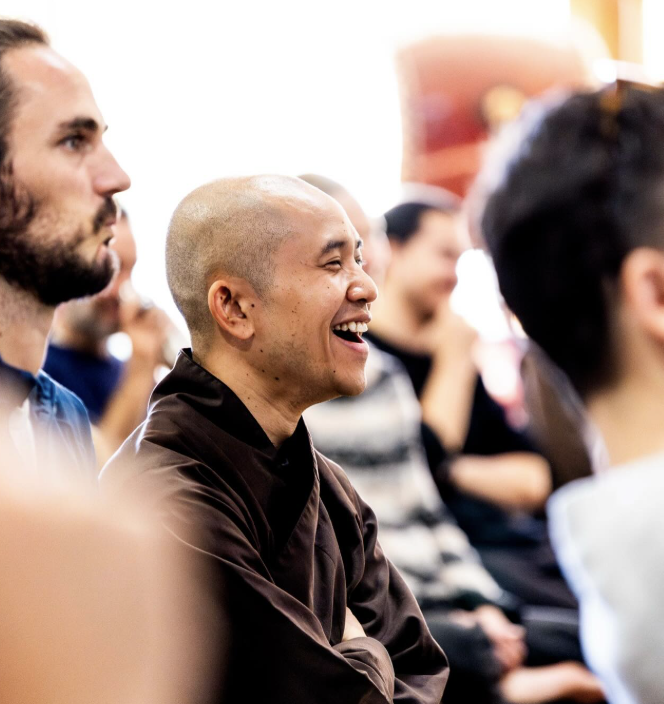 A group of people attending a seminar, with the focus on a smiling, bald man wearing a dark brown jacket.