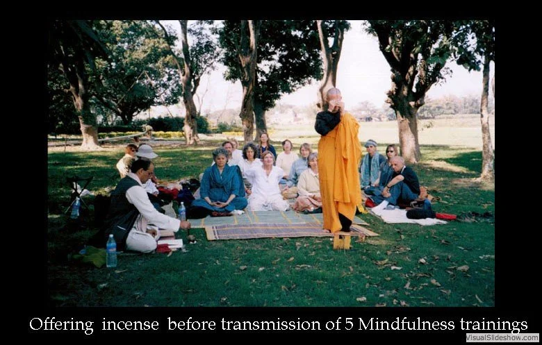 A group of people sitting on mats outdoors under trees, with a monk in orange robes standing in front, offering incense during a mindfulness training session.