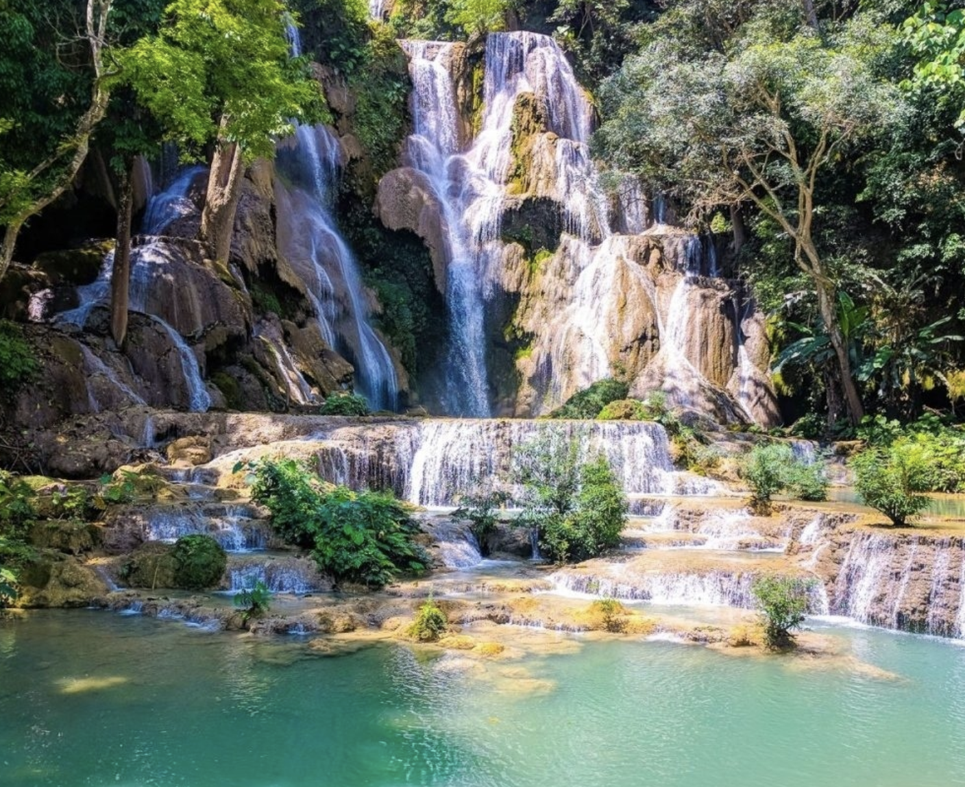 A bustling outdoor market with stalls selling colorful jewelry, bags, clothing, and accessories. Several people are walking and browsing, with some engaged in conversation, under a canopy with hanging lights. Green trees are visible overhead.