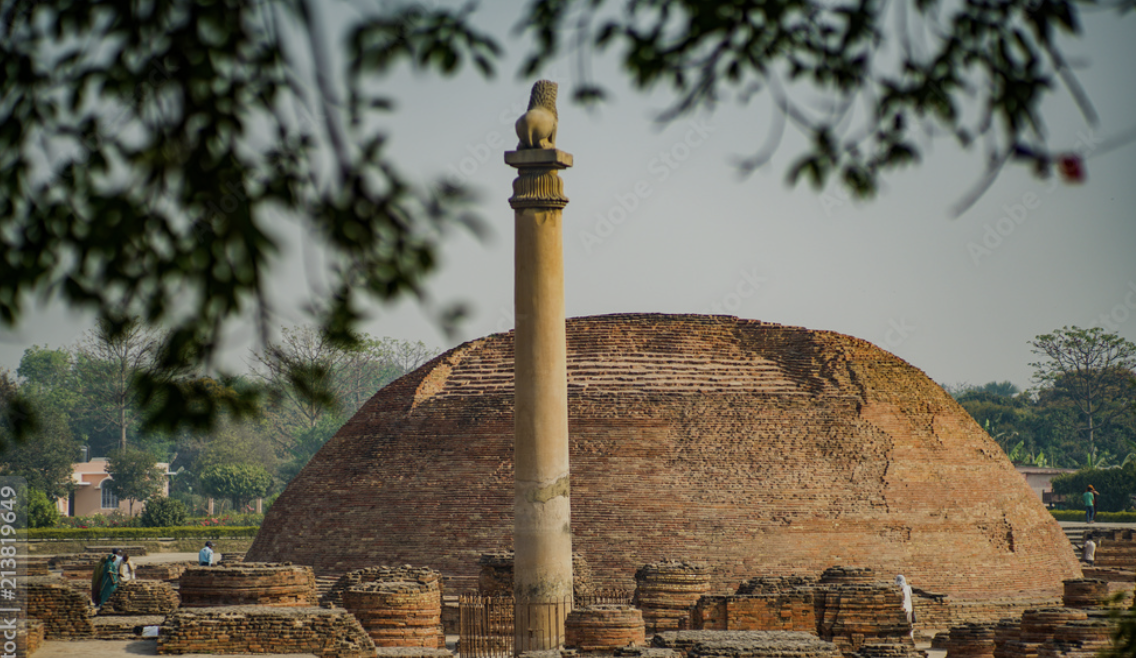 Ancient brick stupa with a single tall pillar topped by a small sculpture, surrounded by trees and ruins.