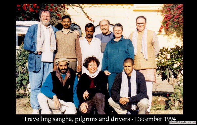 Group of nine diverse people, including monks and pilgrims, posing outdoors in front of flowering bushes, with a man in traditional attire and a cap sitting in the front row, December 1994.