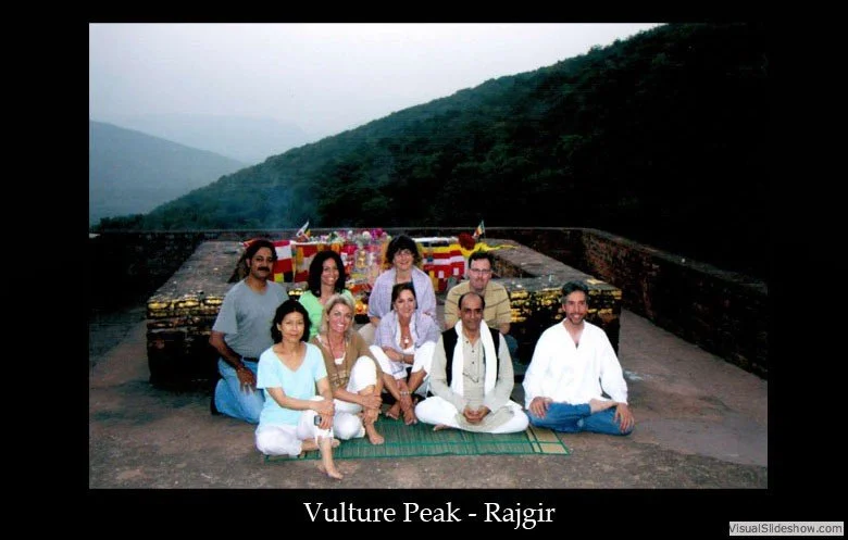 A group of ten people sitting and standing on a green mat outdoors near a decorated platform with flowers and flags, with mountains in the background, during daytime.