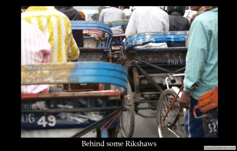A crowded street scene with multiple cycle rickshaws and people walking or riding bicycles, seen from behind.