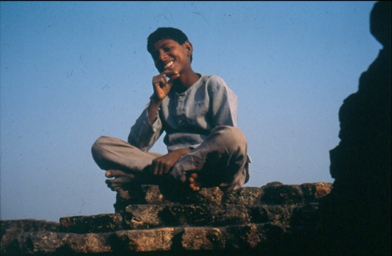 A young boy sitting cross-legged on a brick wall, smiling and touching his chin, against a blue sky background.