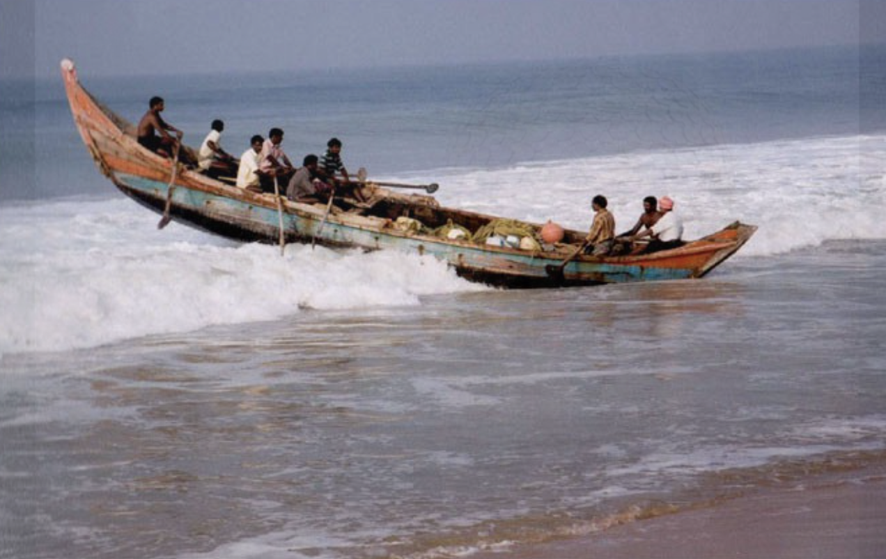 A wooden boat with nine people onboard navigating through ocean waves near the shore.
