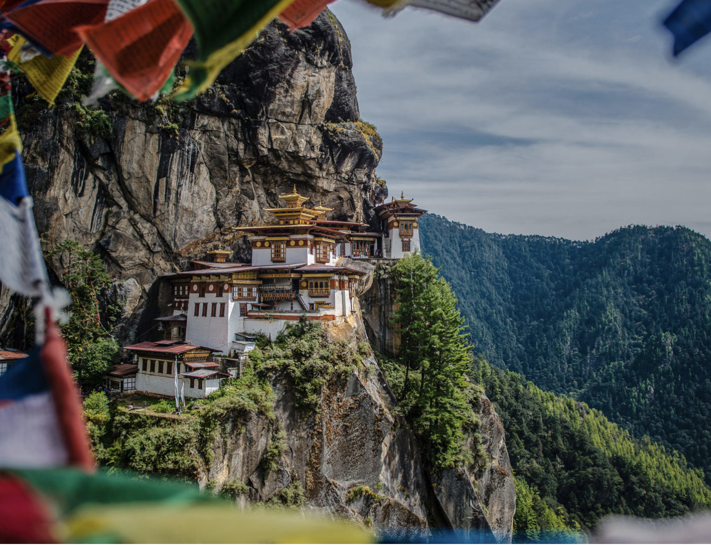 The Tiger's Nest Monastery perched on a cliffside with prayer flags in the foreground, set against a backdrop of forested mountains under a cloudy sky.