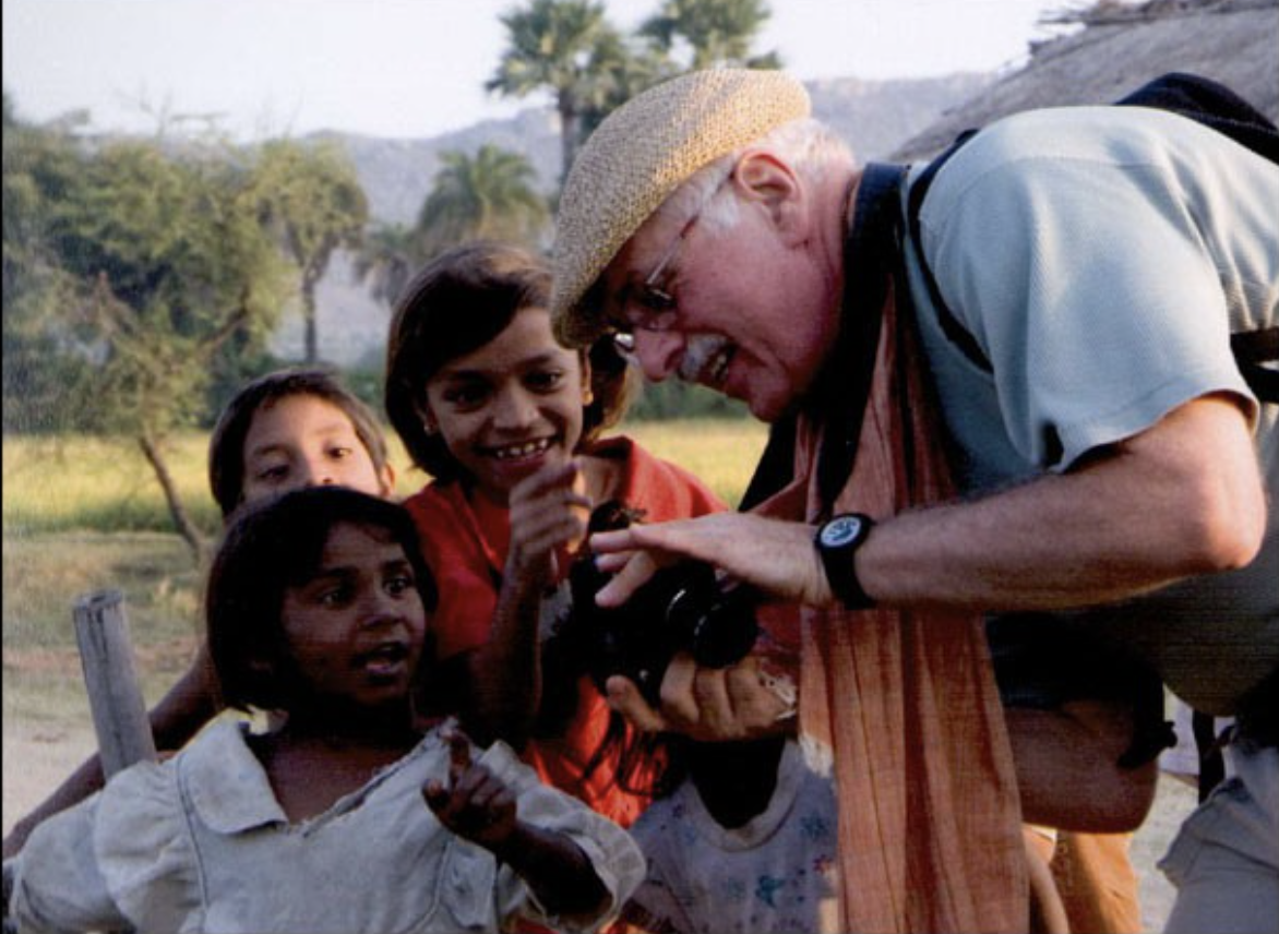 An older man with glasses, a straw hat, and a watch shows a camera to four excited children outdoors with trees and mountains in the background.
