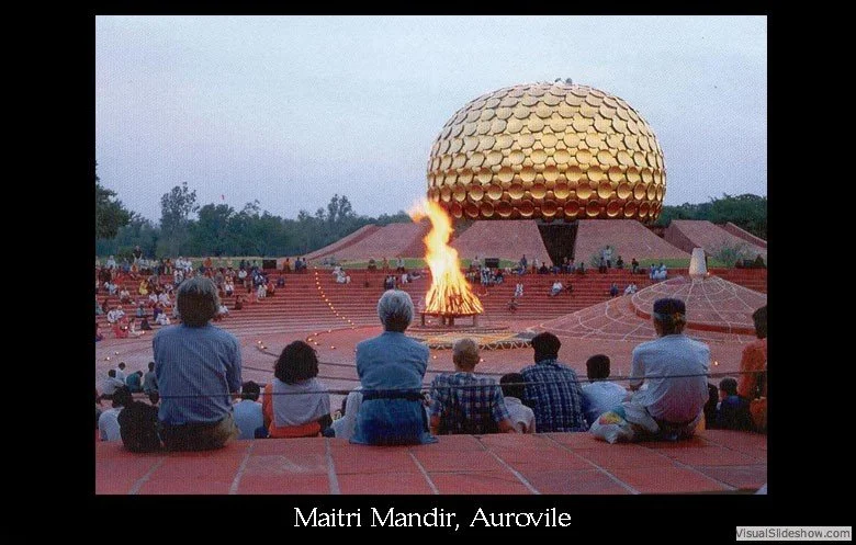 An outdoor gathering at Maitri Mandir in Auroville, with people sitting on steps watching a large, golden, dome-shaped structure with a fire burning in front of it during dusk or evening.
