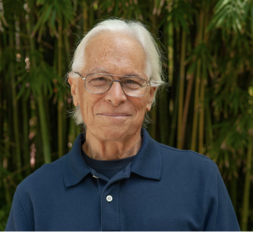 Older man with white hair, glasses, and a slight smile, wearing a navy blue polo shirt, standing outdoors with green palm leaves in the background.