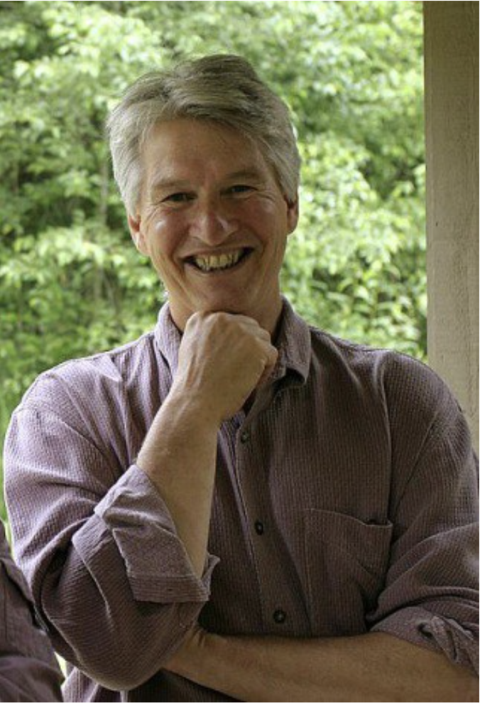 A smiling man with gray hair, wearing a light purple button-up shirt, standing outdoors with green trees in the background.