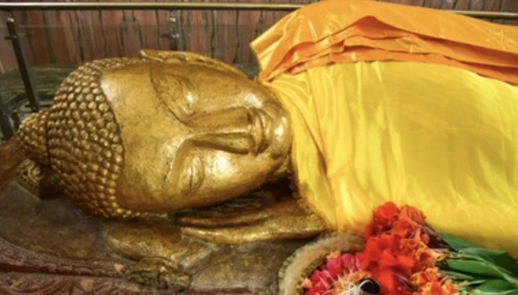 Buddha statue with a golden finish, lying on its side, covered partially with yellow cloth, with flowers nearby.