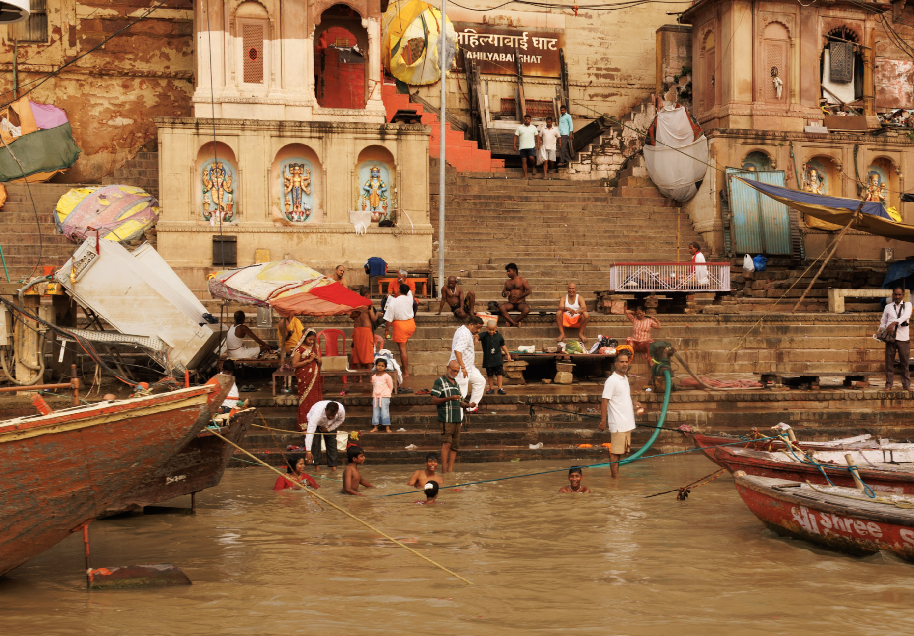 People bathing and gathering on steps along a riverbank with boats docked nearby in India.