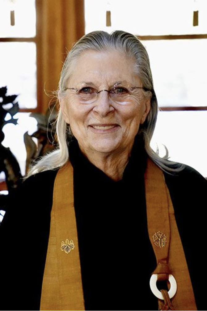 An elderly woman with long gray hair wearing glasses and a black top with a brown stole with gold embroidered symbols, standing indoors near a window.