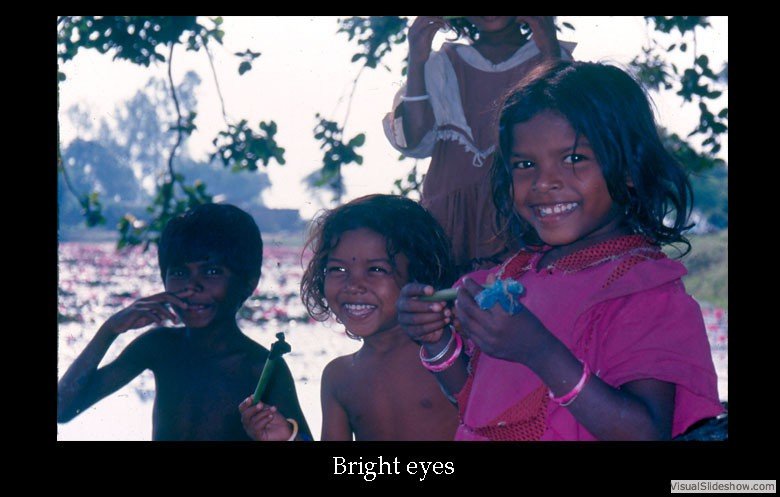 Four children with bright smiles standing outdoors near water and trees, one girl holding a small object, all looking happy.