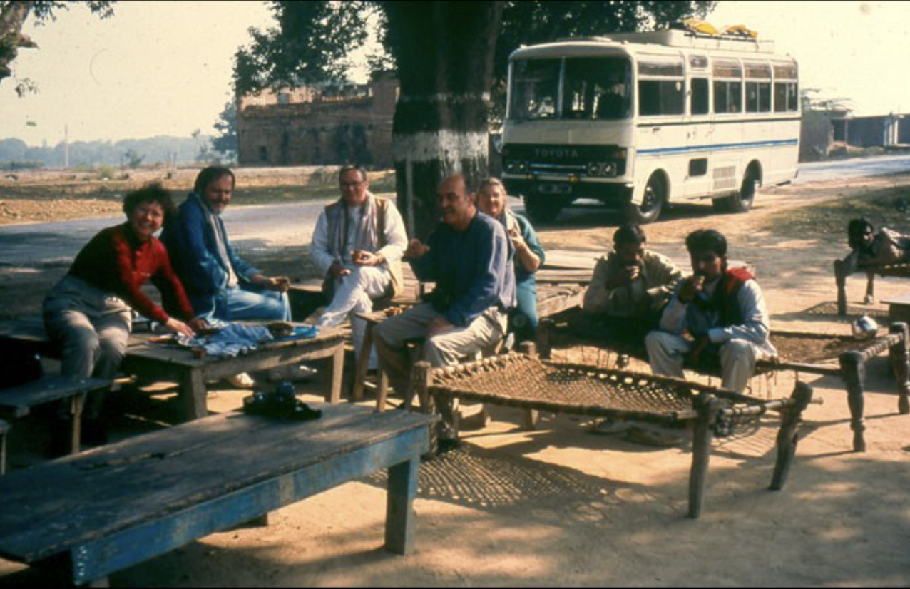 A group of seven men sitting outdoors on benches and wooden tables, with a white Toyota bus in the background on a dirt road in a rural area.
