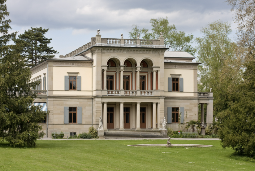 A large, elegant mansion with classical architecture, surrounded by greenery and trees.