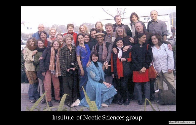 A group of diverse people posing outdoors for a photo, some standing and some kneeling, with a caption "Institute of Noetic Sciences group" at the bottom.