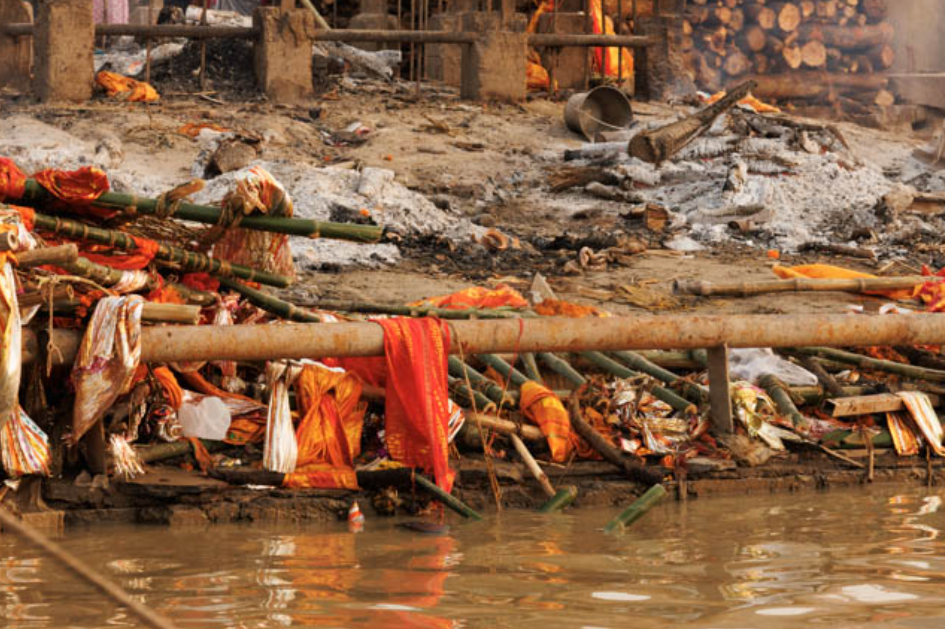 A riverside or lakeside area with makeshift wooden structures, cloth and other items tied to bamboo poles, and a fire burning in the background, with smoky and smoldering debris on the ground.