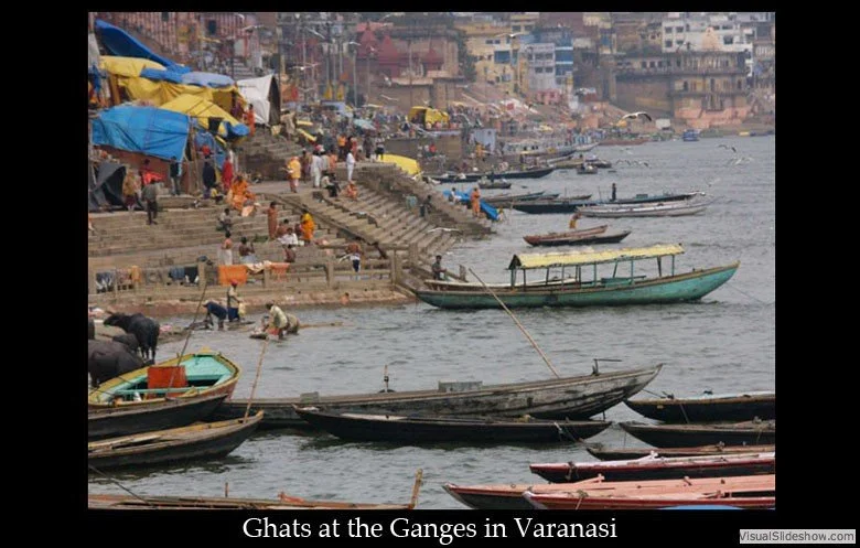 Boats docked along the river and steps leading up to a busy shoreline with tents and people in Varanasi