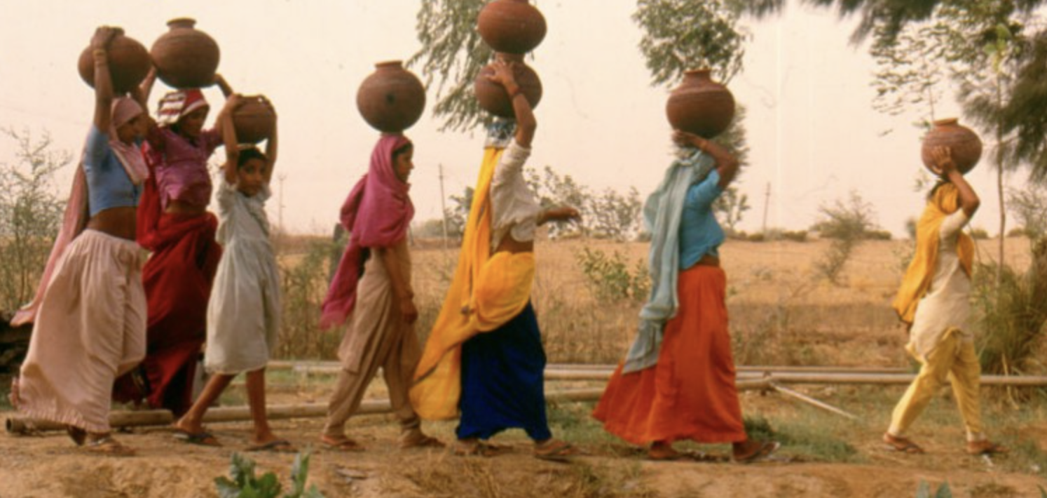 A group of women and children walking outdoors in a line, carrying large clay pots on their heads, in a rural setting with trees and open land.