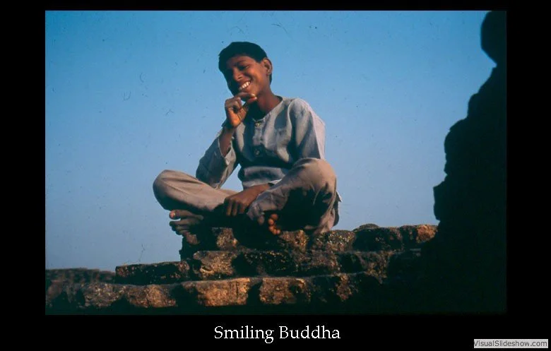 A young person sitting on a rocky surface, smiling and holding a toothbrush in their hand, with a clear blue sky in the background. The caption reads 'Smiling Buddha'.