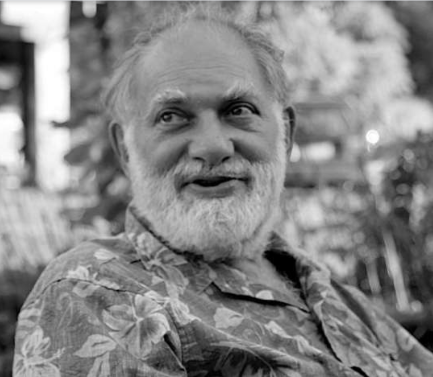 An elderly man with a white beard and mustache, wearing a floral shirt, outdoors in a park or garden setting.