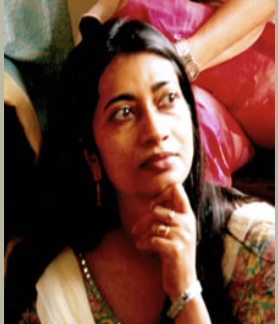 A woman with long dark hair, wearing earrings and a colorful traditional dress, resting her chin on her hand. She appears to be in a thoughtful or attentive mood, with a crowd of people in the background.
