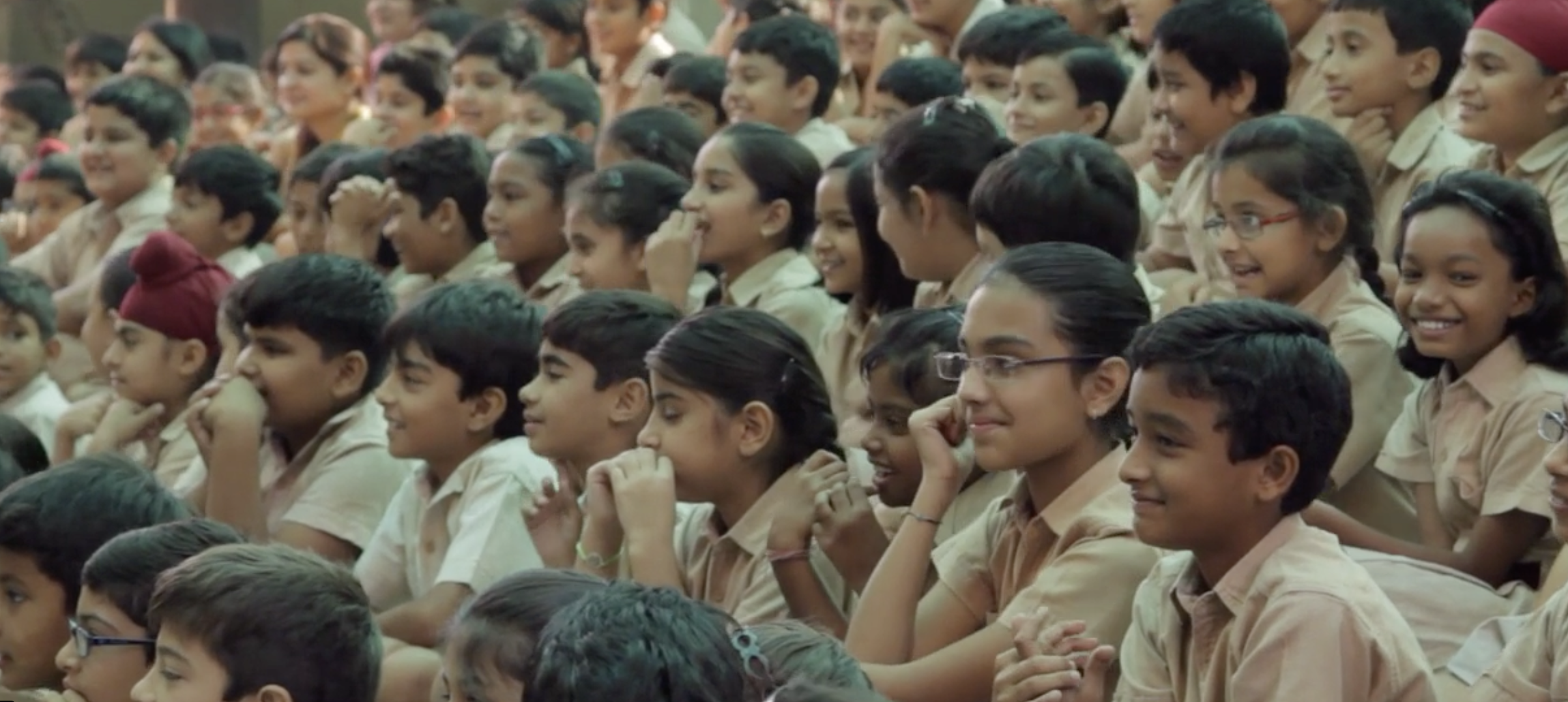 A large group of school children sitting closely together, smiling and laughing during an event or assembly.