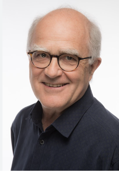 Portrait of an older man with glasses, smiling, wearing a dark shirt against a plain white background.