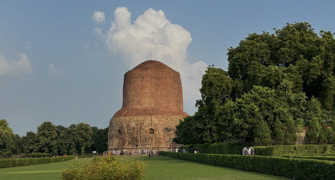 Bing in front of a large, reddish-brown, dome-shaped historic structure on a grassy field with trees and a blue sky with clouds