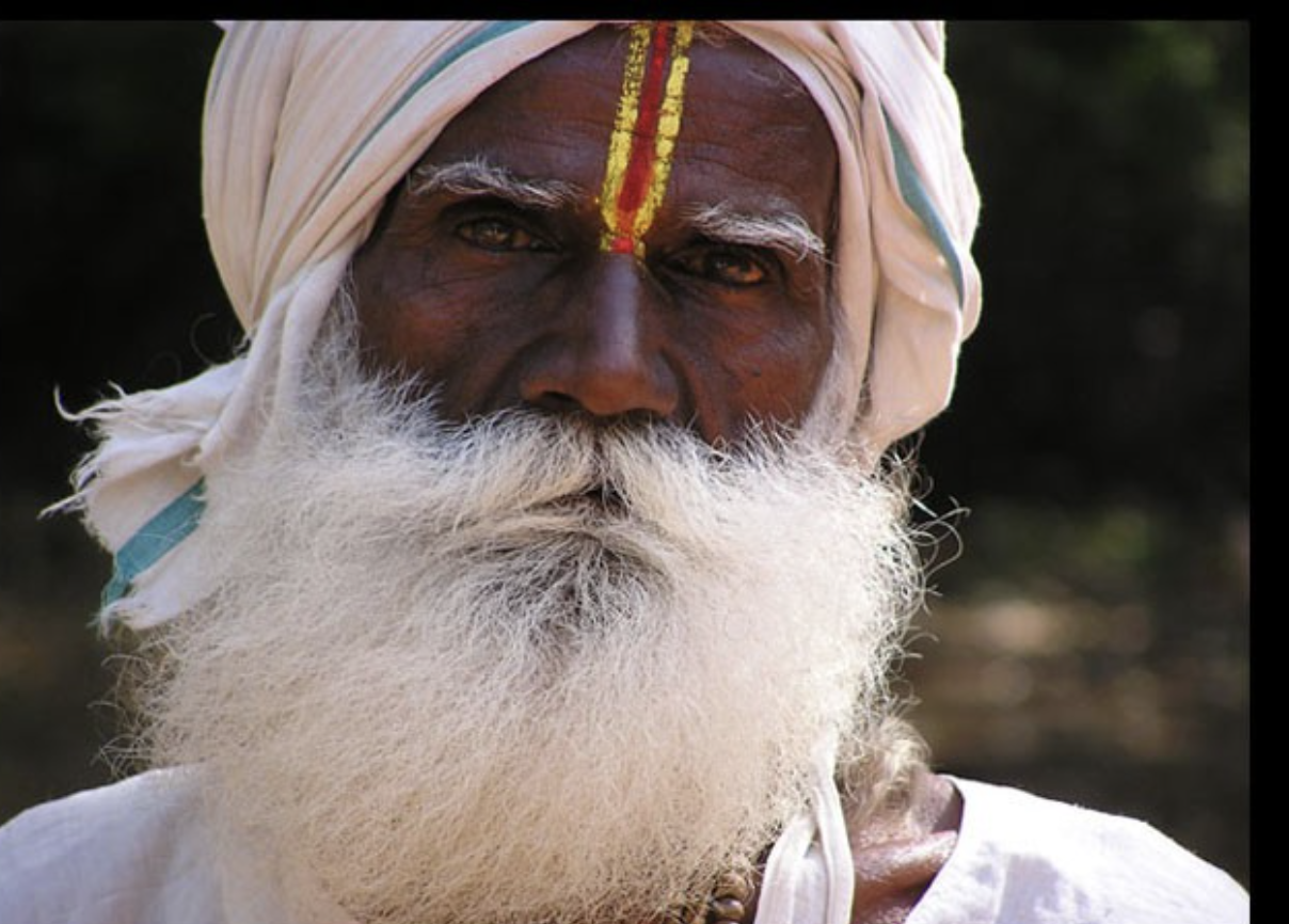 Close-up of an elderly man with dark skin, white beard, and mustache wearing a white turban with red and yellow markings, and a white shirt.