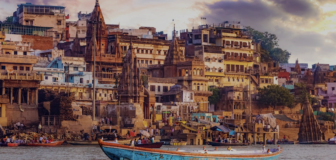 View of a riverside with boats, old buildings, and temples in the background during sunset.