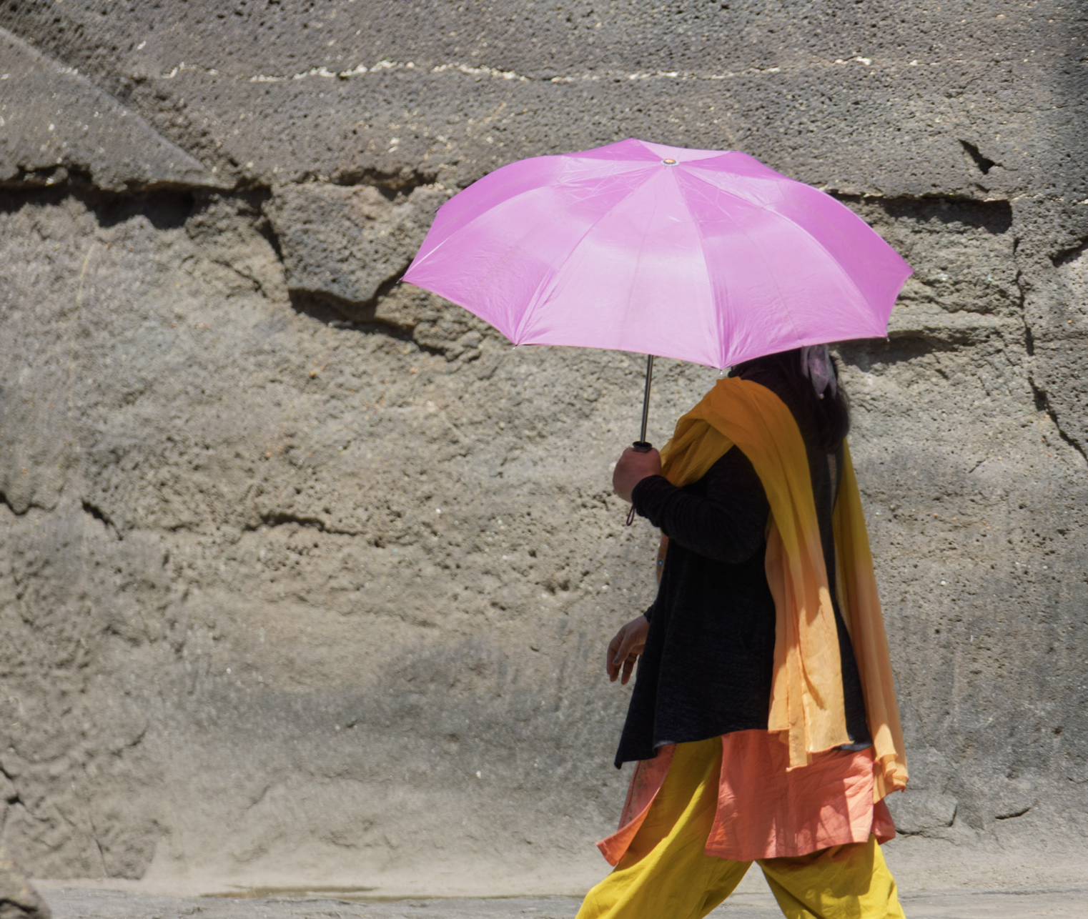 Person walking with a pink umbrella, wearing black top and yellow-orange pants, against a rocky background.