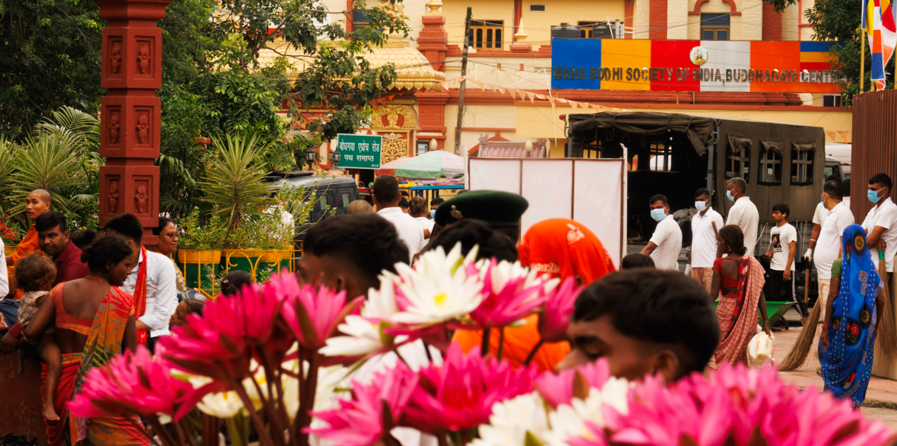 People gathered at a religious or cultural event outside a temple, some wearing traditional attire, with children and adults, and pink flowers in the foreground.