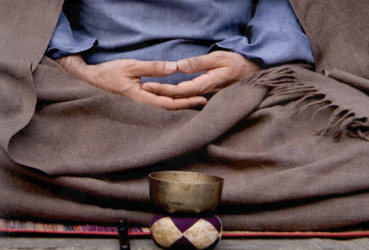 Close-up of a person sitting cross-legged, with hands resting on their lap. They are wearing a blue shirt and brown pants with a fringed shawl. In front of them is a Tibetan singing bowl placed on a decorative cushion.