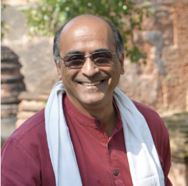 Smiling man wearing sunglasses, a maroon shirt, and a light scarf outdoors with a brick wall and trees in the background.