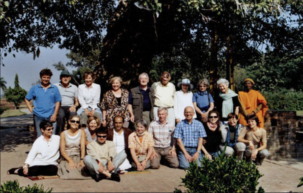 A group of 16 people posing outdoors under a large tree on a sunny day, with some people sitting on the ground and others standing behind them. They are smiling and dressed casually.