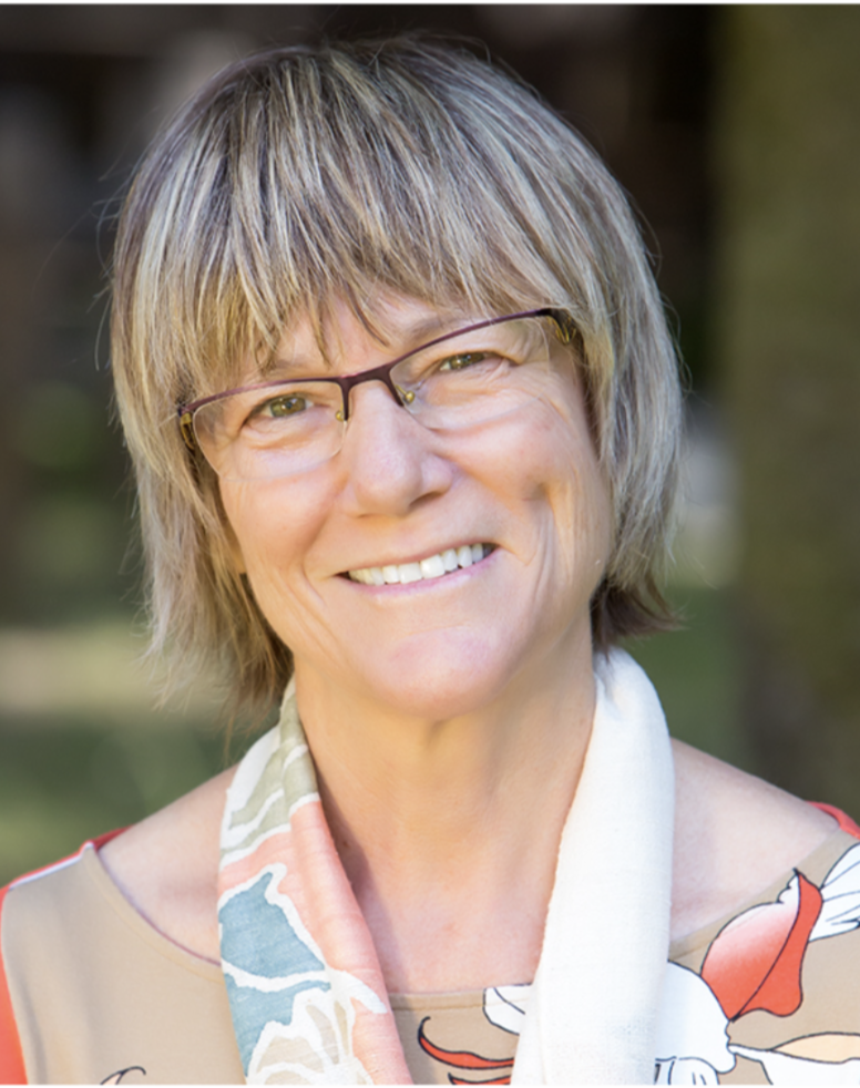 A smiling middle-aged woman with short, light brown hair, glasses, and wearing a colorful scarf and patterned top outdoors.