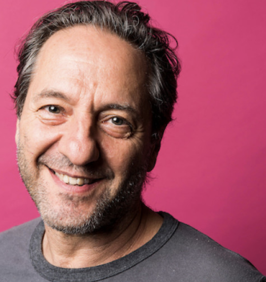 Close-up of smiling middle-aged man with gray hair and beard against a pink background.