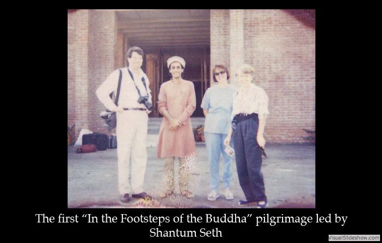 Historical photo of four people standing outside a brick building, with a caption about the first 'In the Footsteps of the Buddha' pilgrimage led by Shantum Seth.