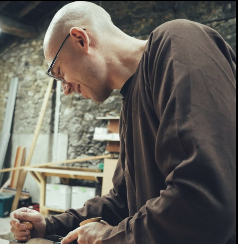 Smiling man wearing glasses, a red shirt, and a white scarf outdoors with a blurred background of trees and brick walls.