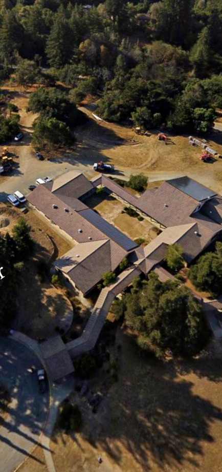 Aerial view of a large residential property with multiple connected buildings, trees, and a dirt parking area with several parked cars.