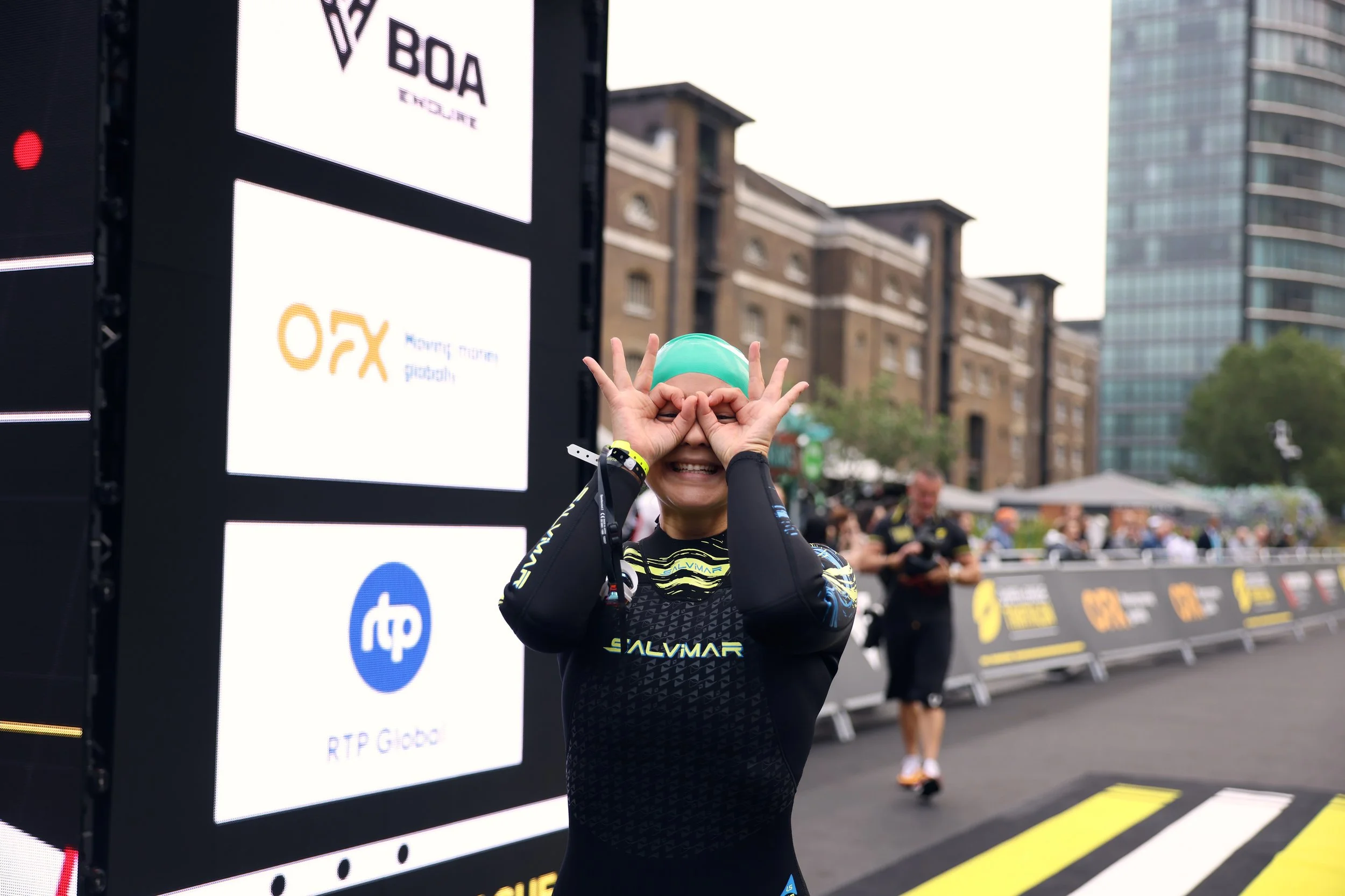 A woman in a black compression suit and teal swim cap smiling and making goggles with her hands at an outdoor sporting event, with banners and other people in the background.