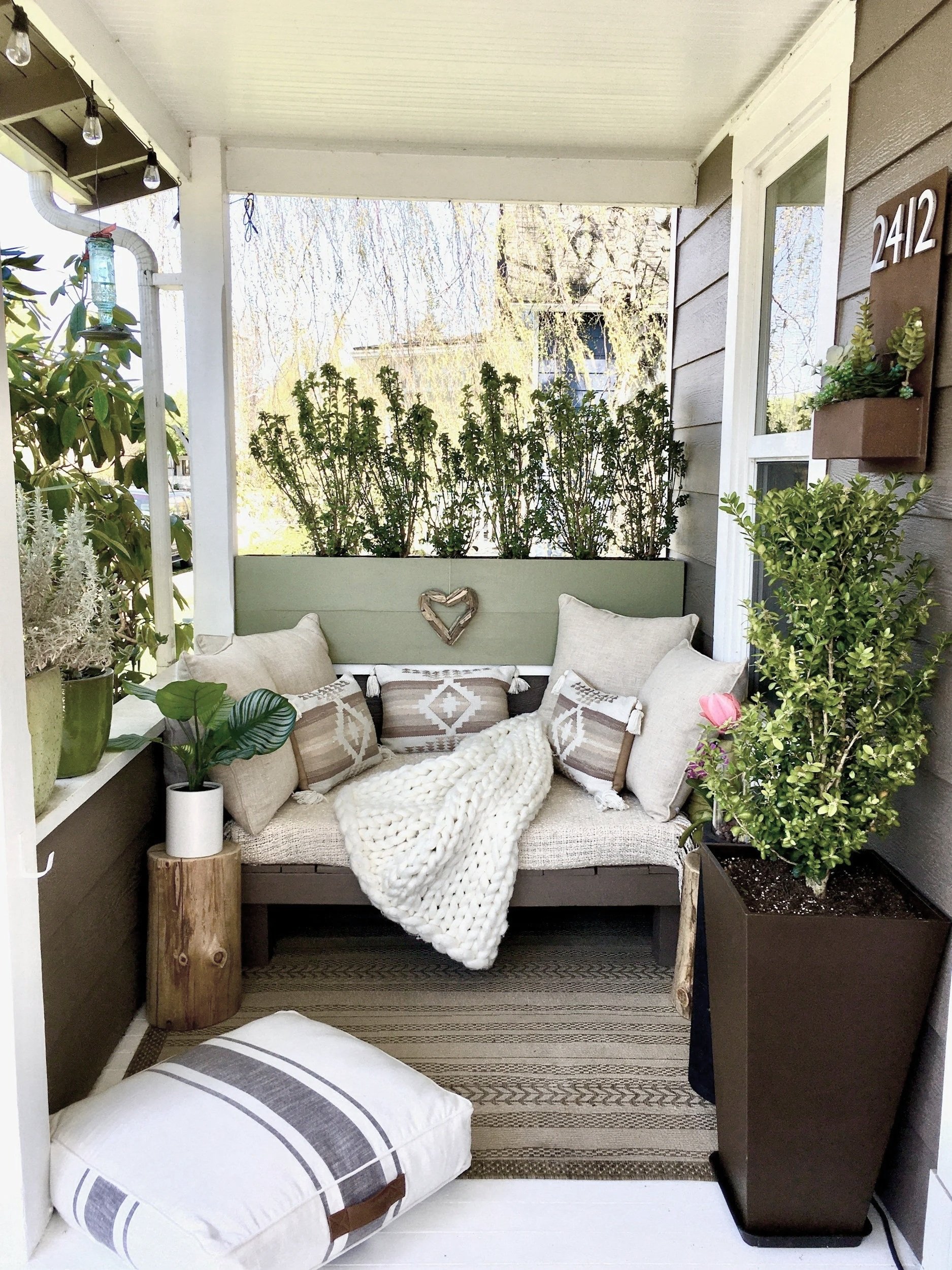 Cozy enclosed porch with a cushioned bench, surrounded by plants, pillows, knitted throw, and decorative elements.