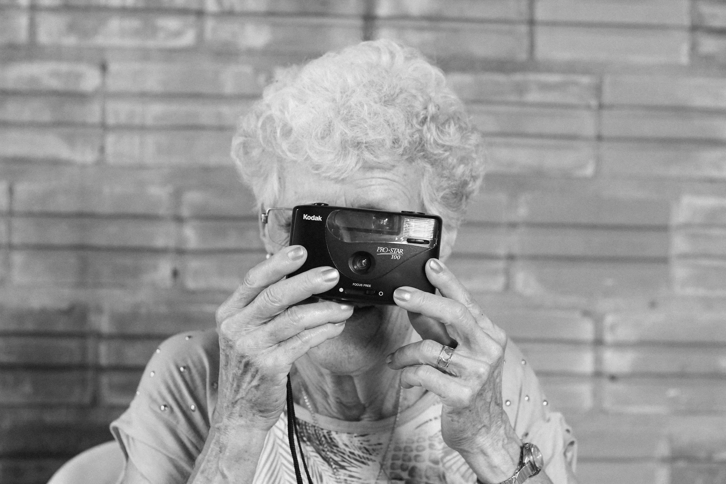 An elderly woman with curly white hair taking a photograph with a film camera against a brick wall background.
