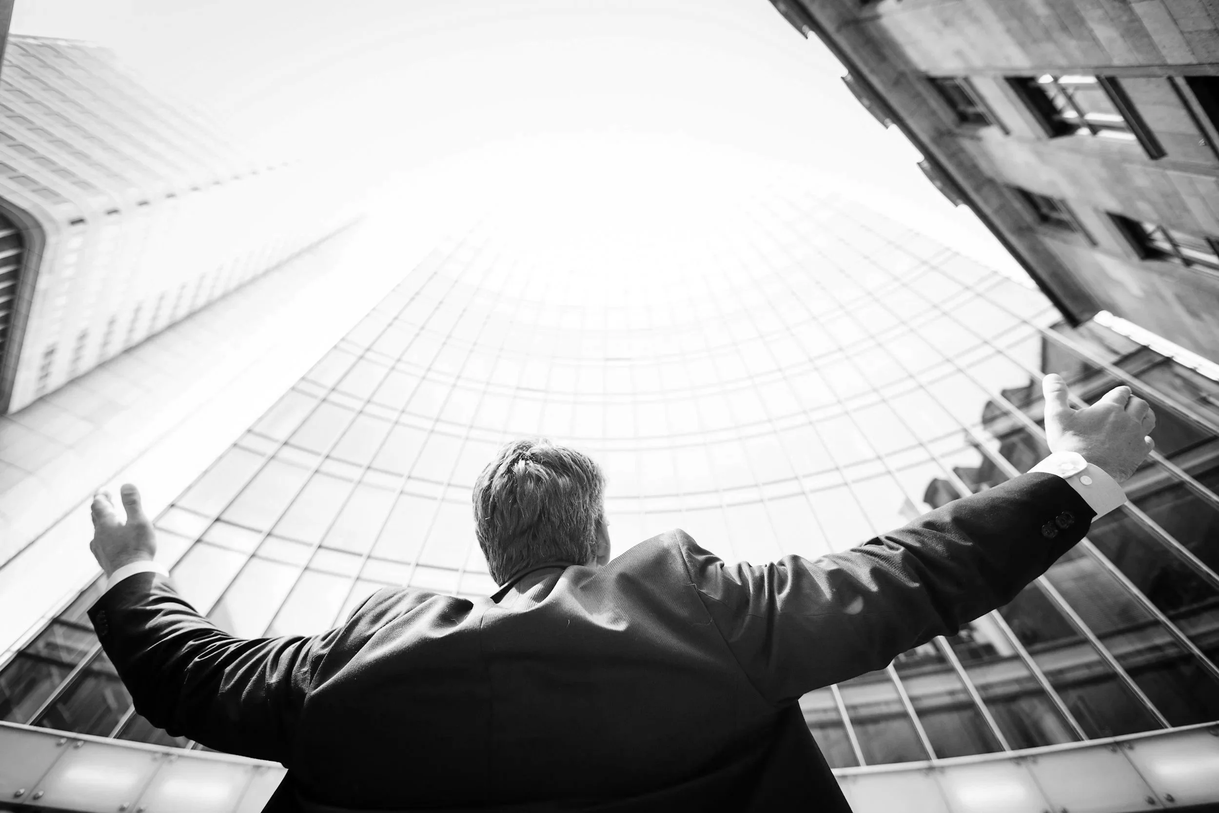 Businessman in a suit standing with arms outstretched in front of a tall, glass skyscraper, looking upward.
